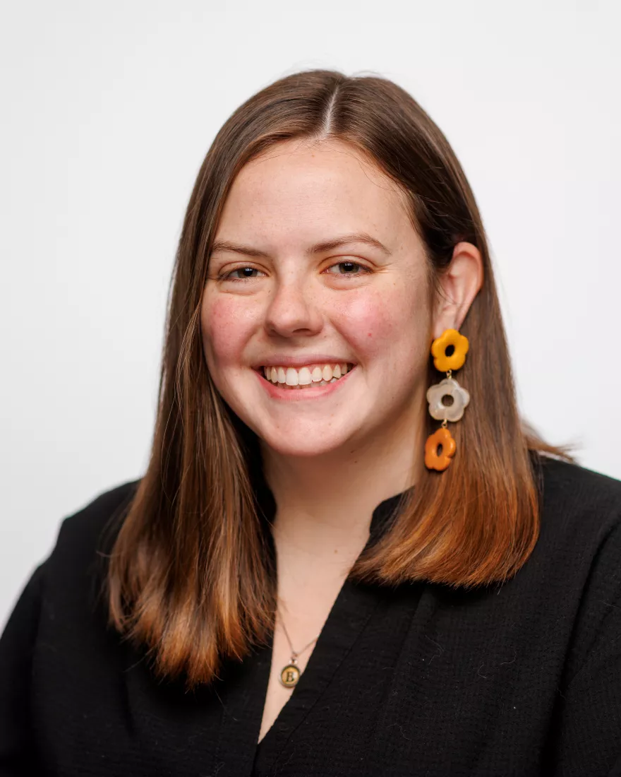 A woman with brown hair poses for a headshot in front of a white background. She is wearing a black blouse, a necklace with a B typewriter key pendant, and earrings with three orange flowers.